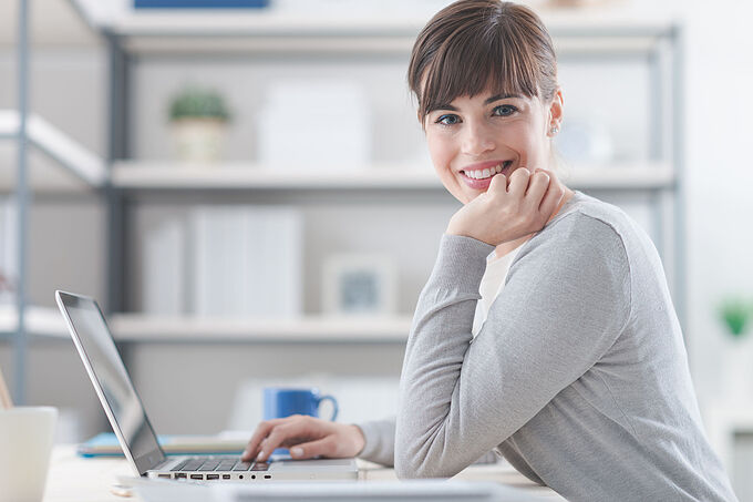 61531086 - happy confident businesswoman sitting at office desk and working with a laptop, she is smiling at camera